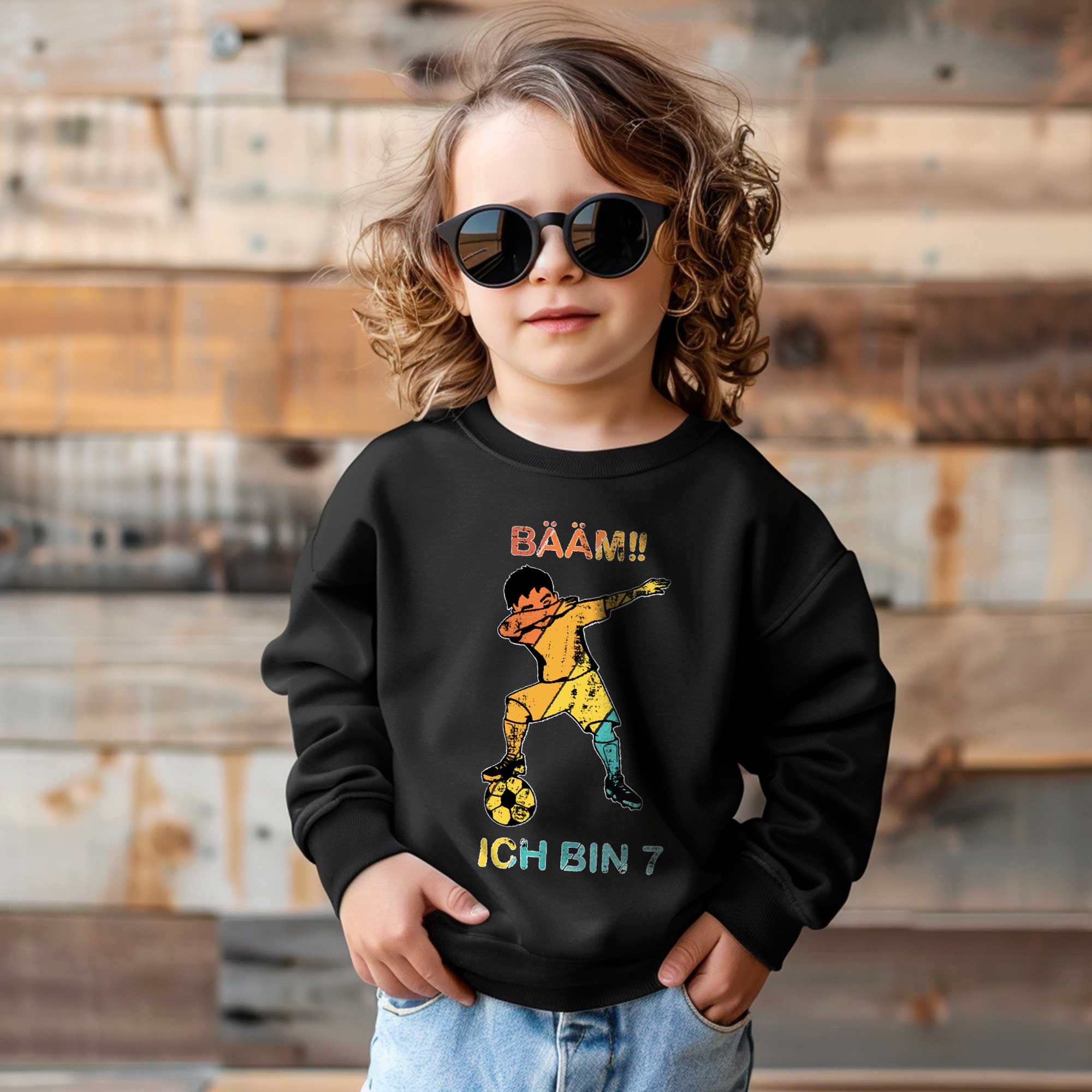 Kind mit Sonnenbrille trägt schwarzes Sweatshirt mit bunter Fußballgrafik und Spruch ‚BÄÄM!! Ich bin 7‘ – stylisches Geburtstags-Outfit für Jungen.
