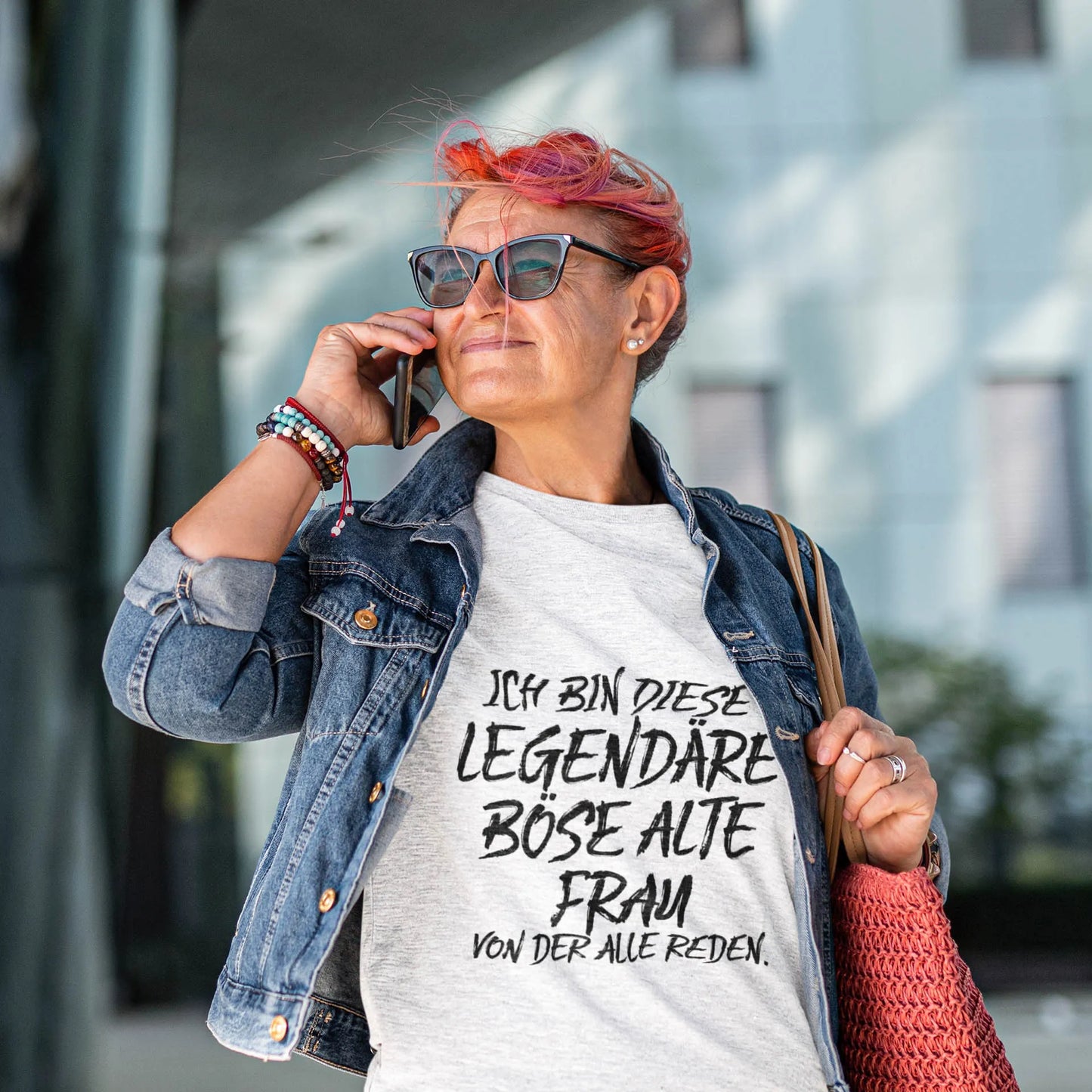 Selbstbewusste Frau mit Sonnenbrille und Jeansjacke telefoniert und trägt ein graues Statement-T-Shirt mit Aufdruck „Ich bin diese legendäre böse alte Frau, von der alle reden“ – moderne Damenmode mit Humor.